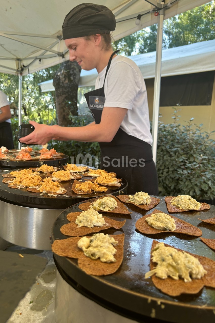 Une cuisinière prépare des galettes garnies sur des plaques chauffantes lors d'un événement en extérieur.