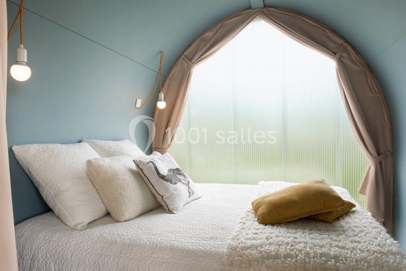 Lit double avec coussins et couverture dans une chambre aux murs bleus, éclairée par une grande fenêtre avec rideaux beiges.