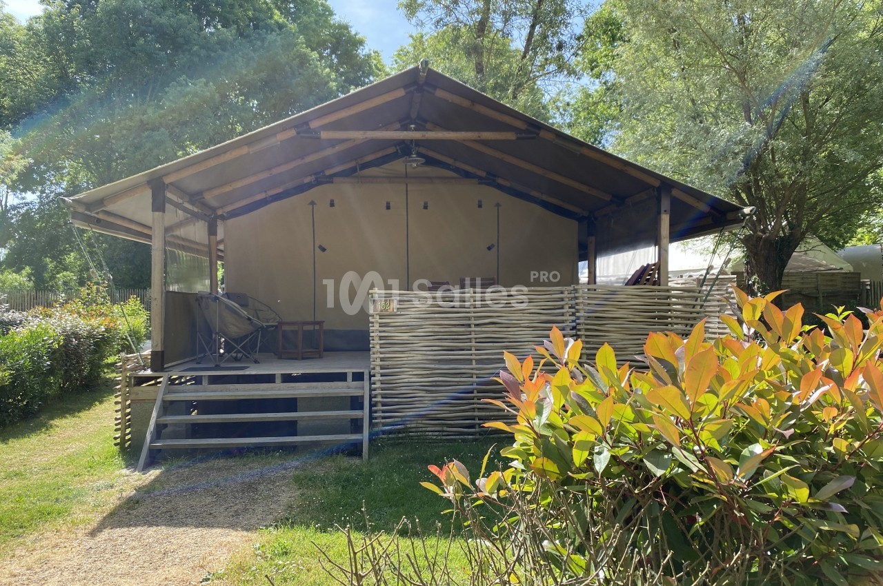 Tente lodge en toile avec terrasse en bois, entourée de végétation sous un ciel ensoleillé.