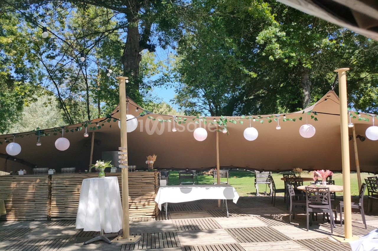 Espace extérieur aménagé avec tables, chaises et guirlandes lumineuses sous une tente beige, entouré de verdure.