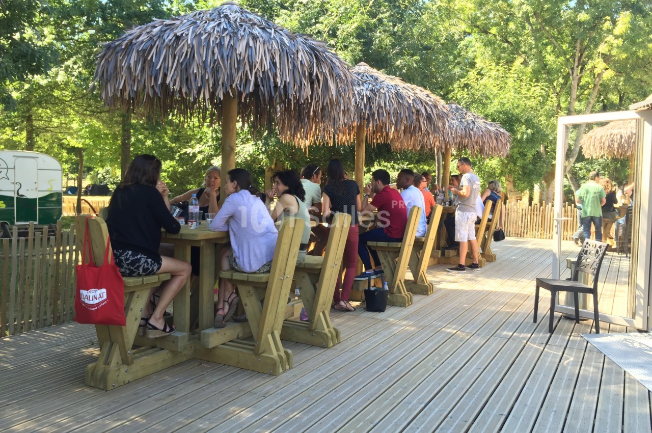 Des personnes assises et debout autour de tables en bois sous des parasols en chaume sur une terrasse ensoleillée.