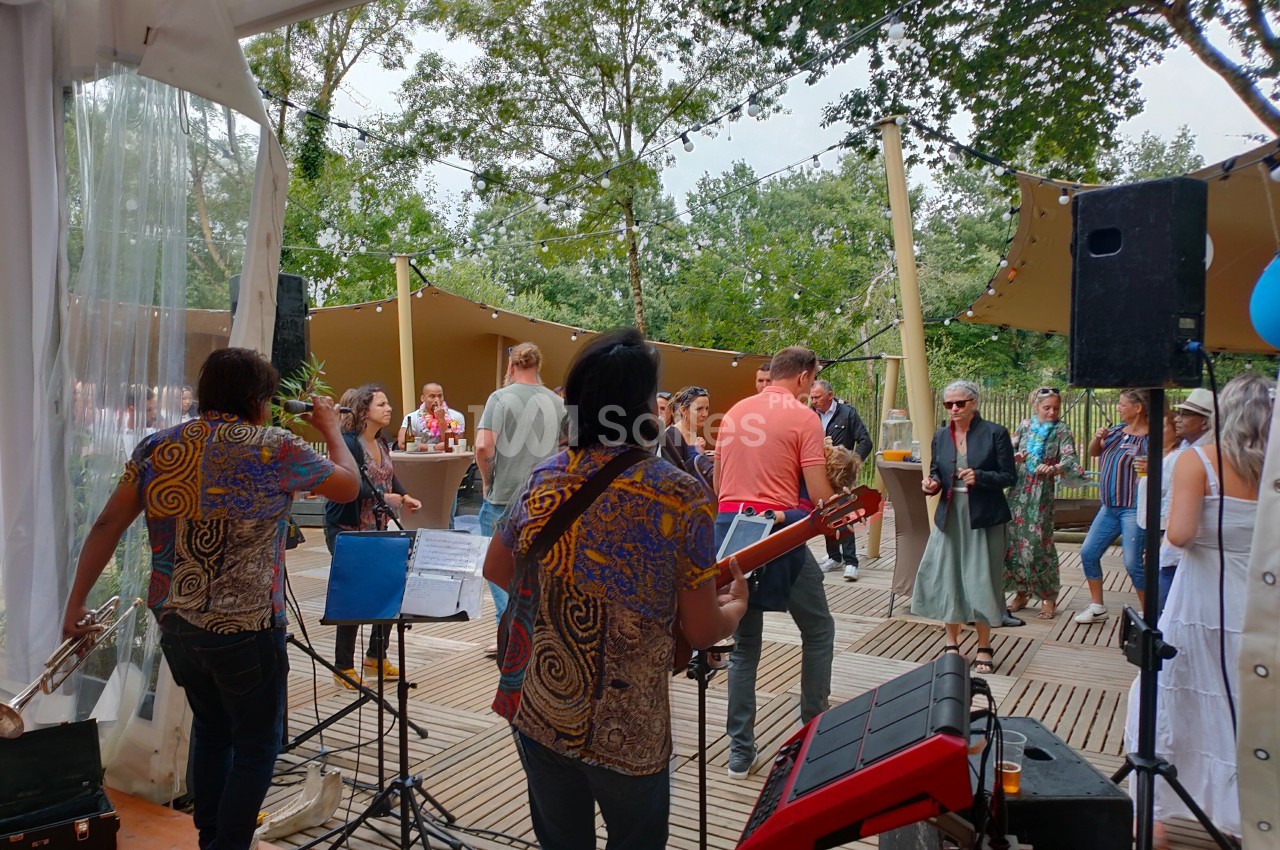 Groupe de musiciens jouant en plein air devant un public dans un espace boisé avec des décorations suspendues.