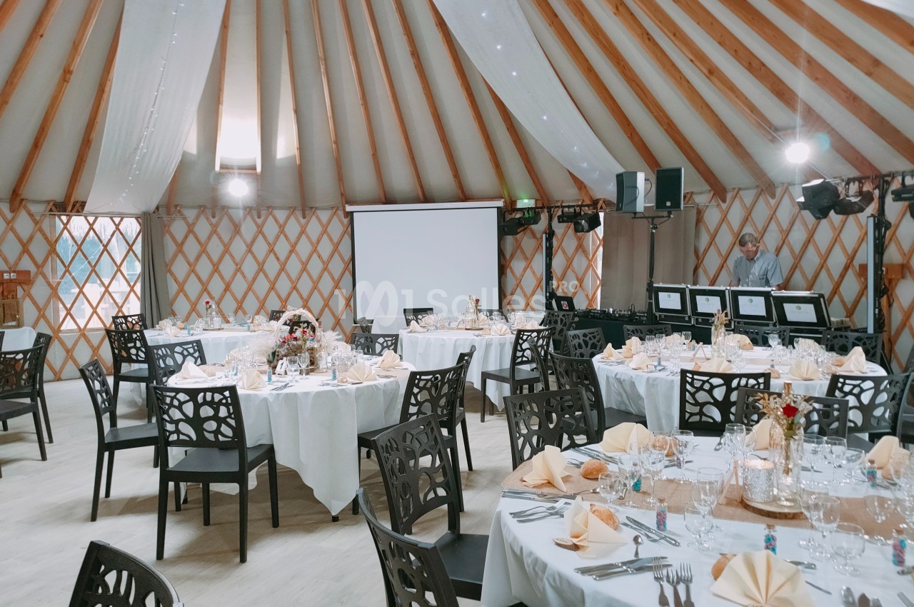 Salle de réception décorée avec des tables rondes dressées, nappes blanches et chaises noires, sous une structure en bois.