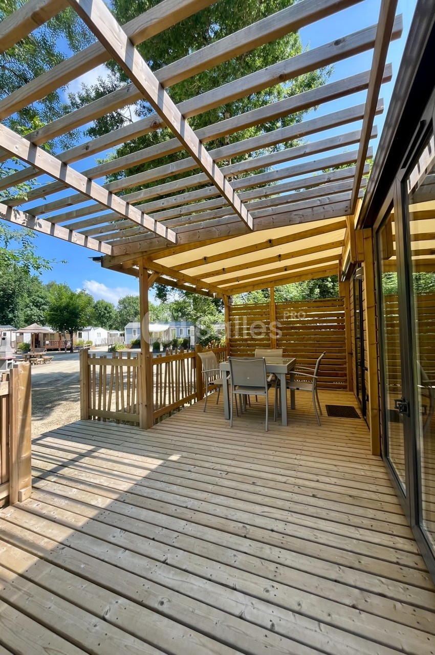 Terrasse en bois avec pergola, table et chaises, entourée de mobil-homes dans un cadre arboré.