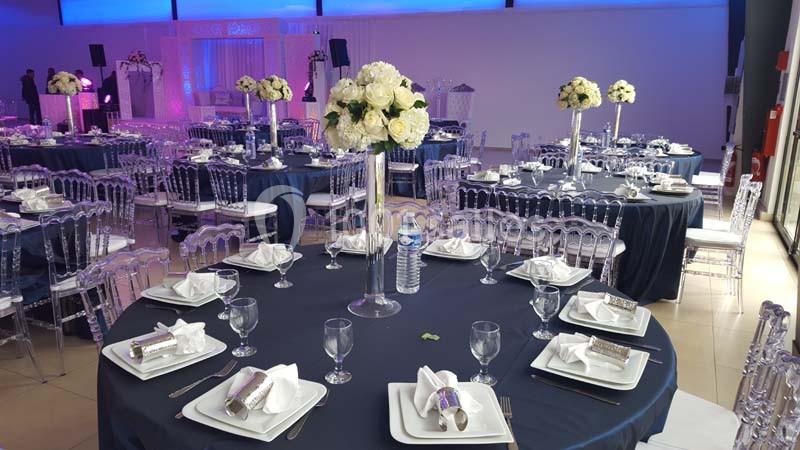 Salle de réception décorée avec des tables rondes nappées de bleu, ornées de centres de table floraux et de vaisselle…