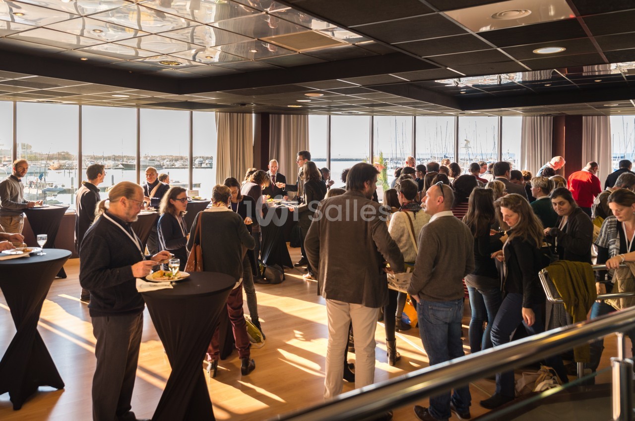 Groupe de personnes échangeant debout autour de tables hautes dans une salle lumineuse avec vue sur un port.