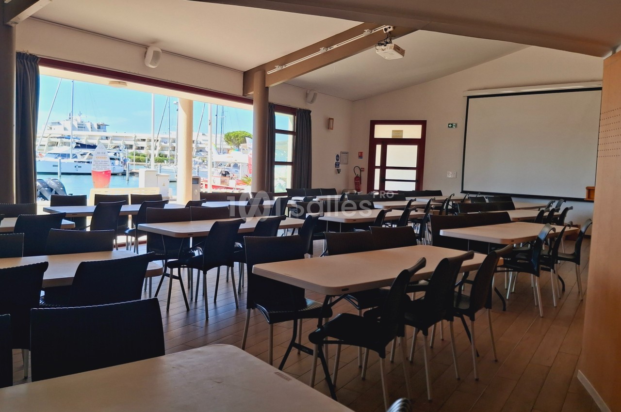 Salle de réunion lumineuse avec des tables et chaises disposées face à un écran, vue sur un port à travers les fenêtres.