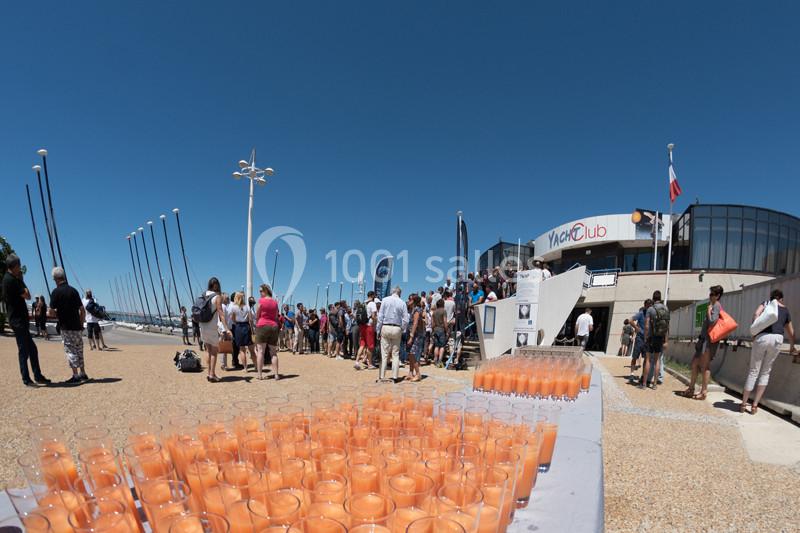 Des verres de jus d'orange alignés sur une table en extérieur, avec une foule rassemblée devant un bâtiment moderne.