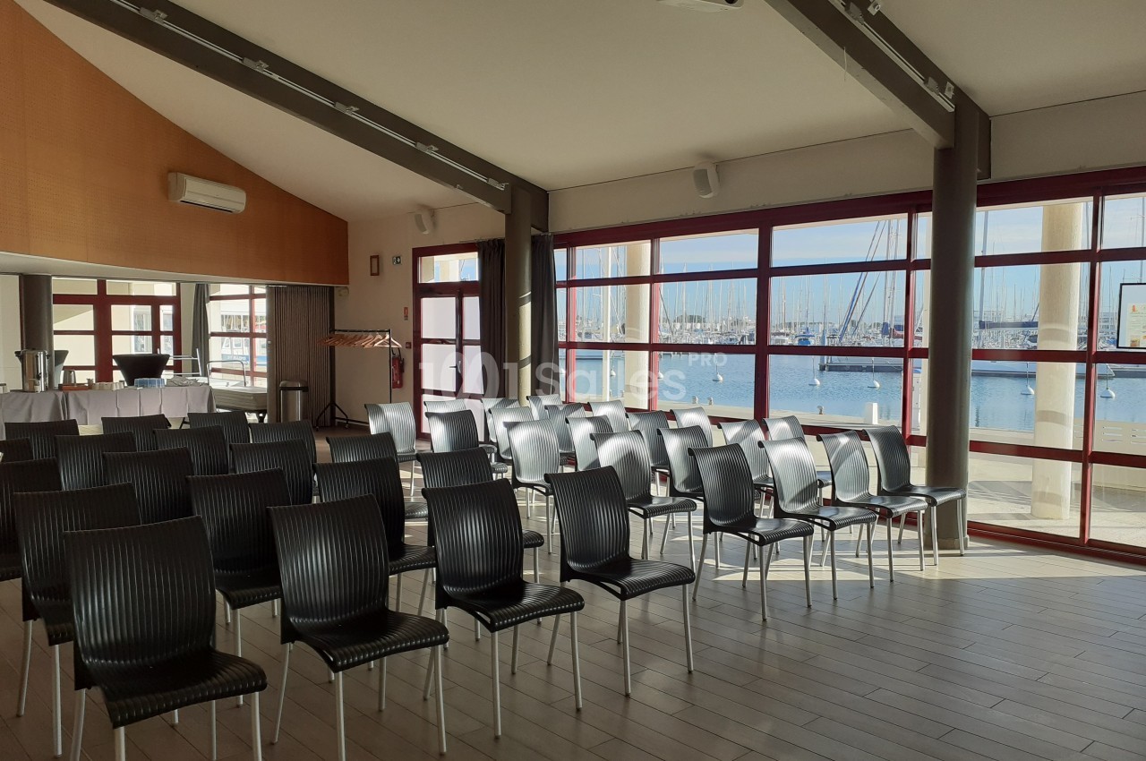 Salle lumineuse avec des rangées de chaises noires vides, grandes baies vitrées donnant sur un port de plaisance.