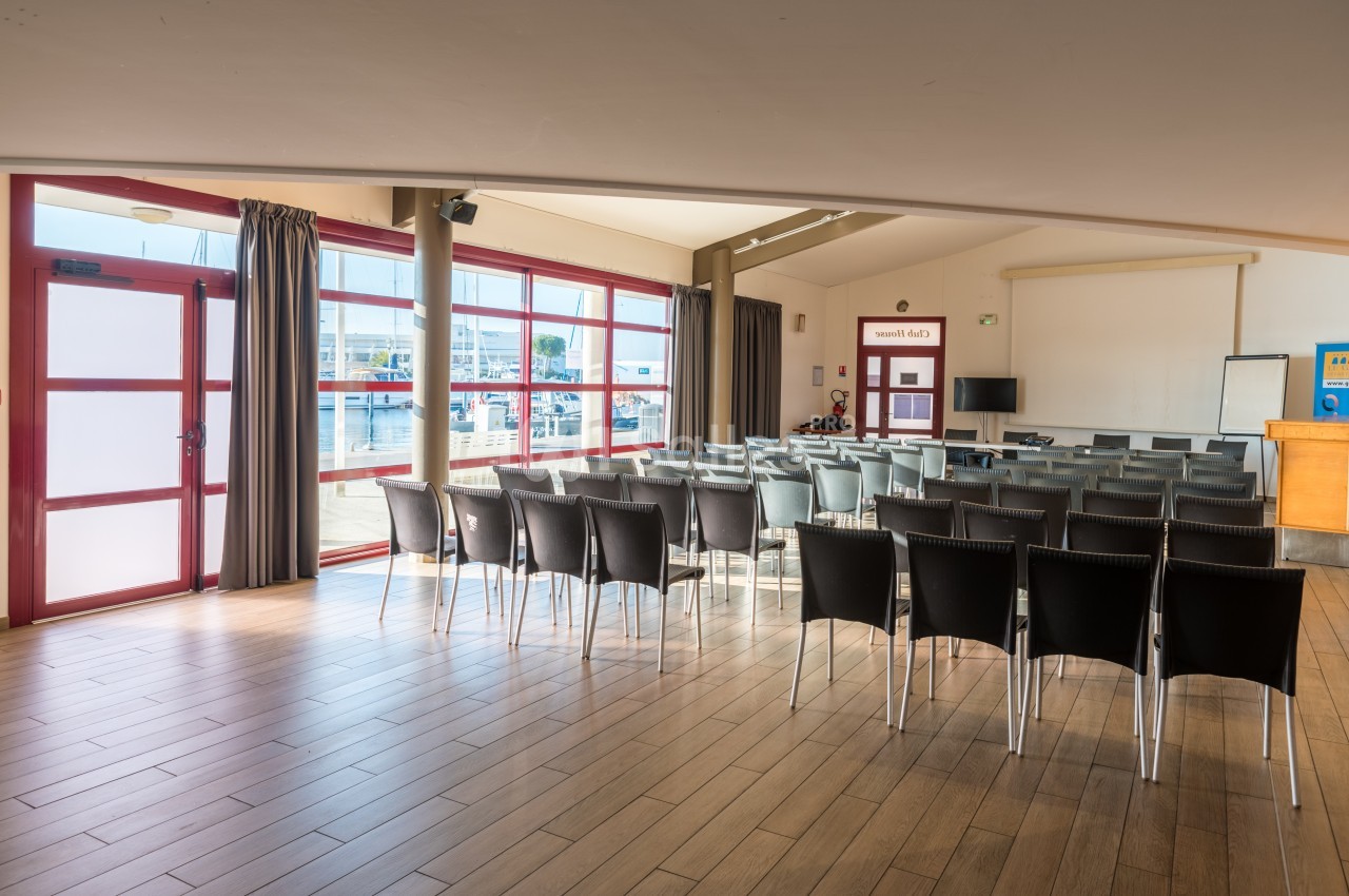 Salle de réunion lumineuse avec des chaises alignées, un écran de projection et de grandes fenêtres donnant sur un port.