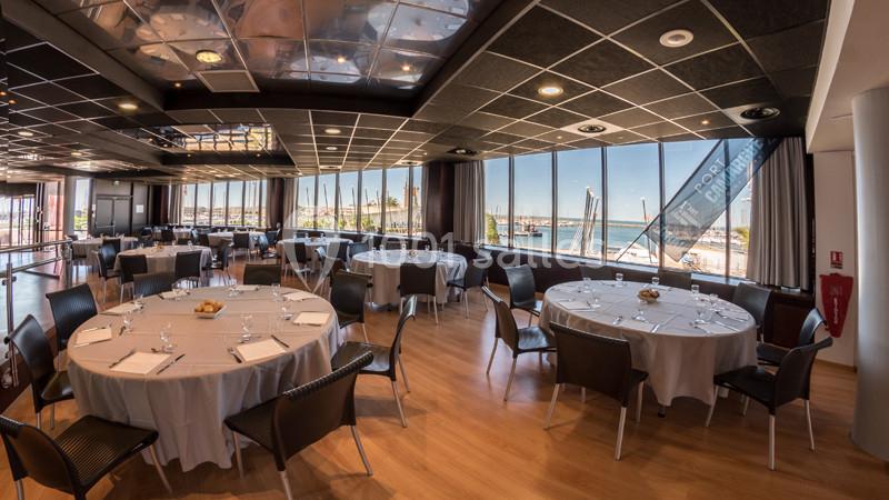 Salle de réception lumineuse avec tables rondes dressées, grandes baies vitrées offrant une vue sur un port.
