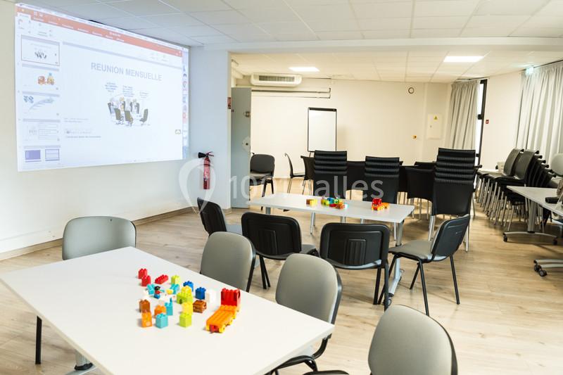 Salle de réunion lumineuse avec tables, chaises, écran de projection et blocs de construction colorés sur les tables.