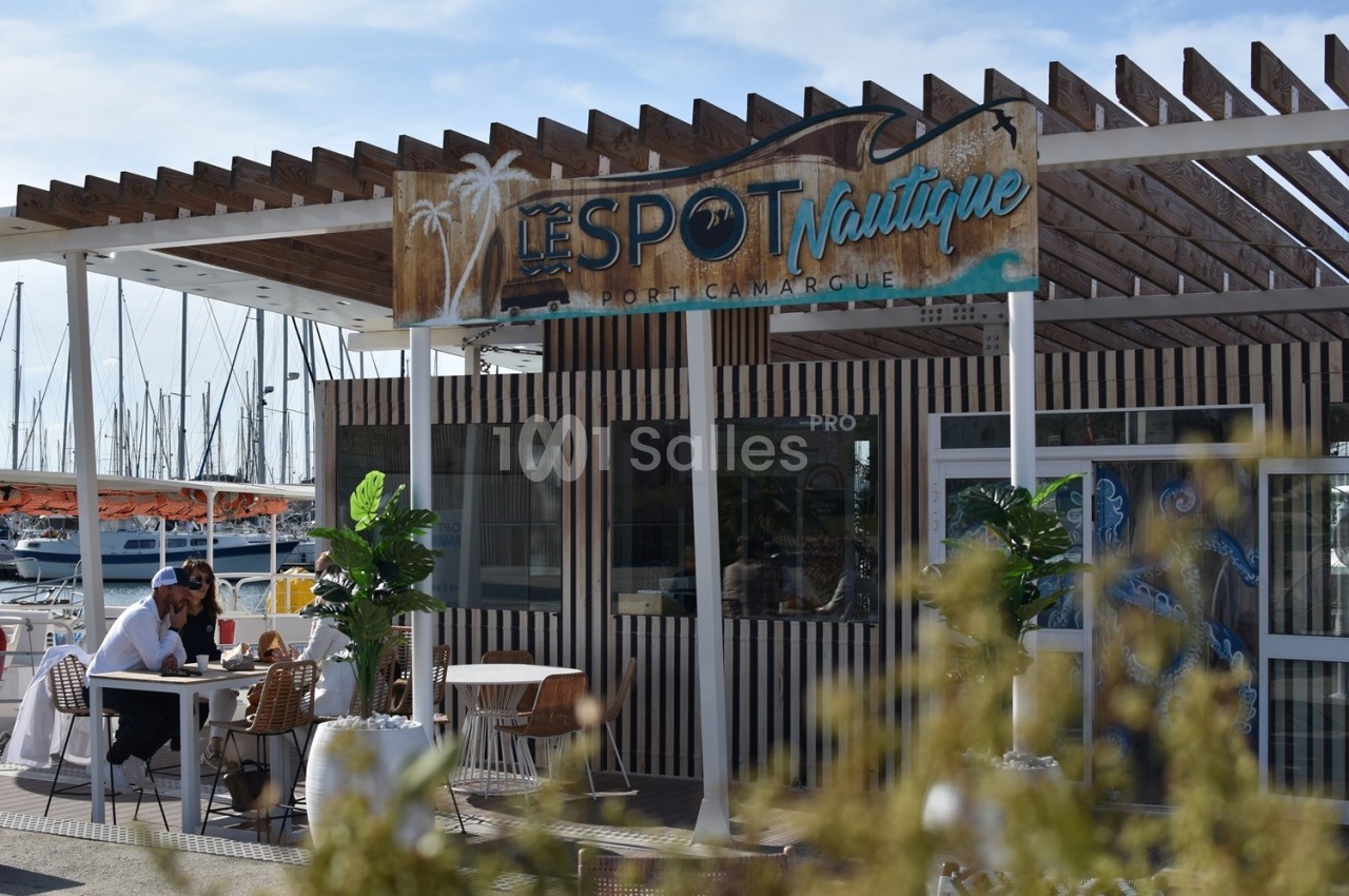 Terrasse d'un restaurant en bois avec vue sur un port de plaisance, tables occupées et végétation en premier plan.