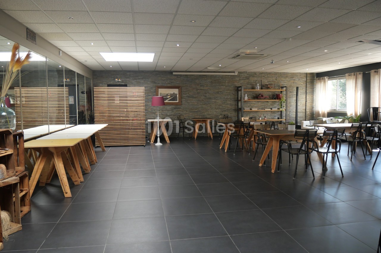 Salle lumineuse avec tables en bois, chaises noires, sol en carrelage sombre et murs en pierre.