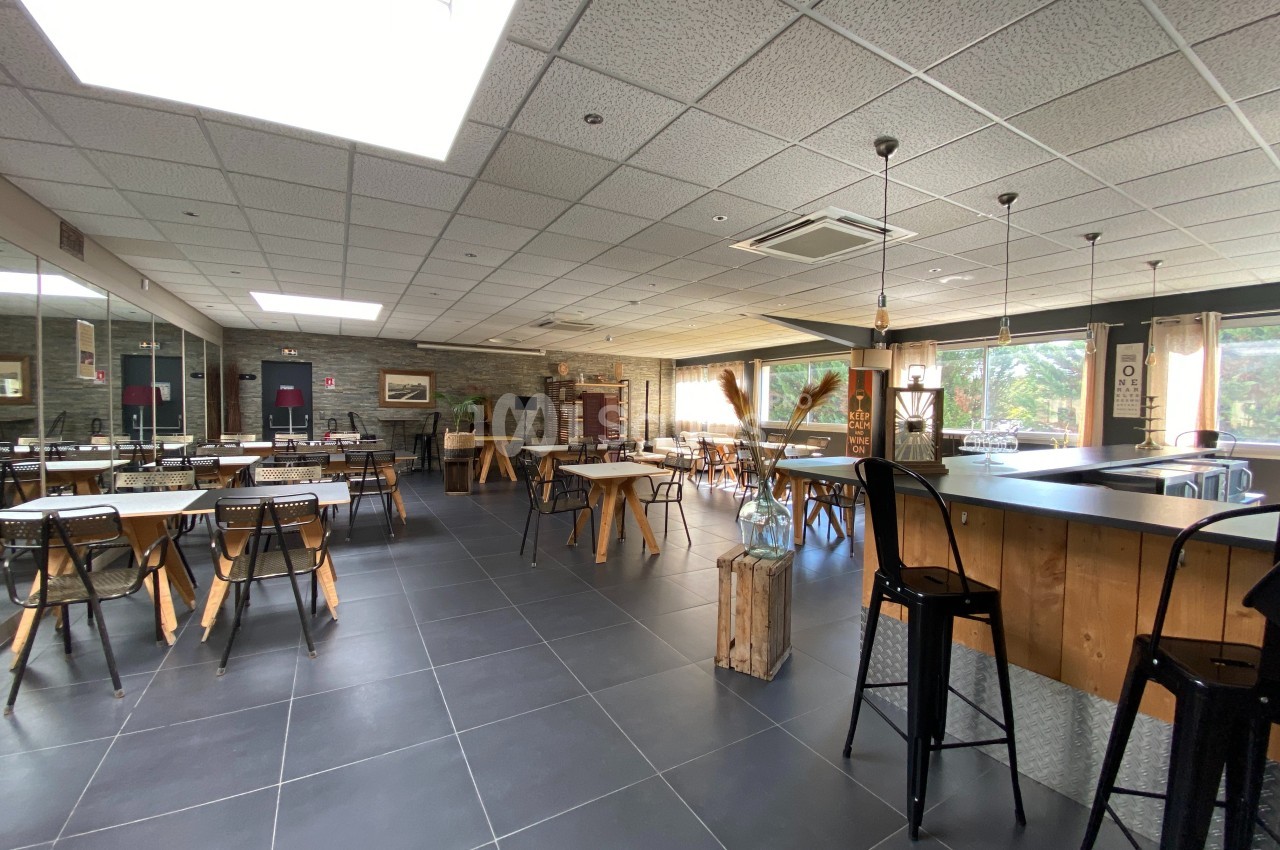 Salle lumineuse avec tables, chaises, bar en bois et grandes fenêtres donnant sur un espace extérieur verdoyant.