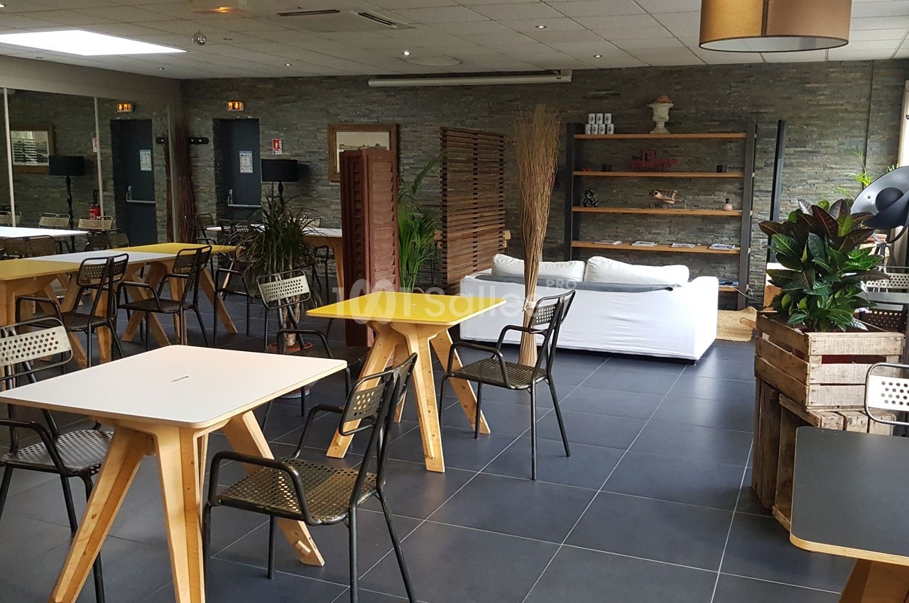 Salle lumineuse avec tables en bois, chaises noires, canapé blanc, étagères décoratives et plantes vertes.