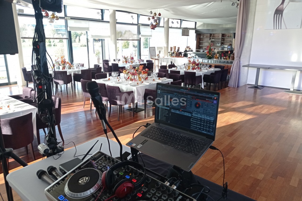 Poste de DJ avec platines et ordinateur en premier plan, salle de réception décorée et dressée en arrière-plan.