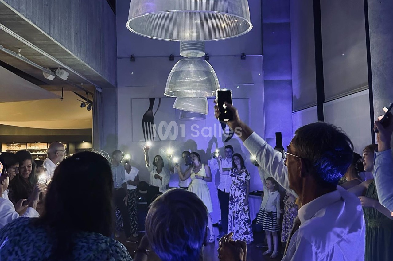 Des personnes rassemblées dans une salle sombre, éclairées par des lumières de téléphones et des lampes suspendues.