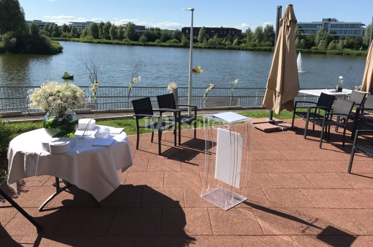 Terrasse ensoleillée avec vue sur un lac, tables dressées et parasols fermés, cadre calme et verdoyant.