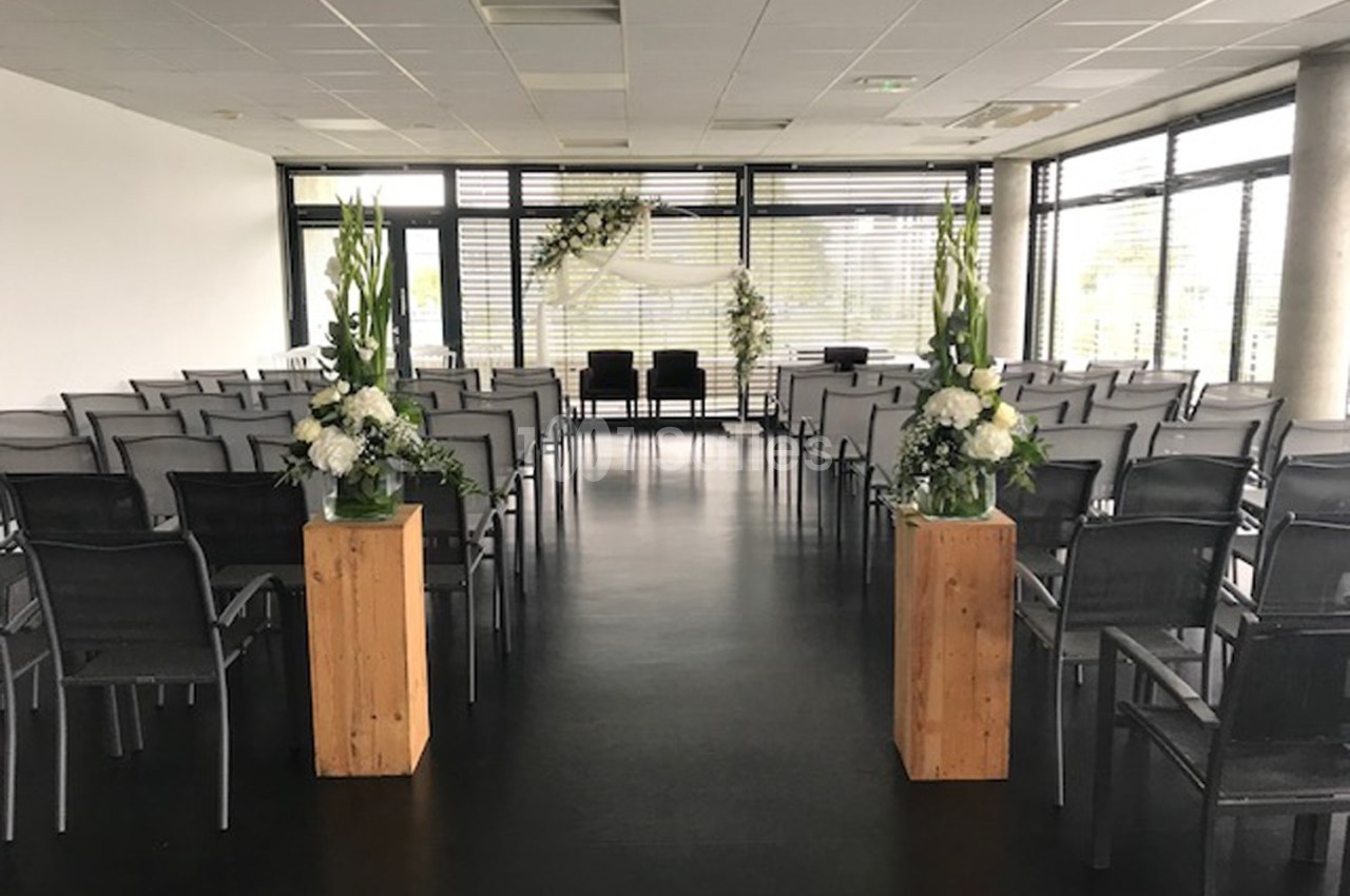 Salle de cérémonie lumineuse avec des rangées de chaises noires et des décorations florales blanches sur des supports en…
