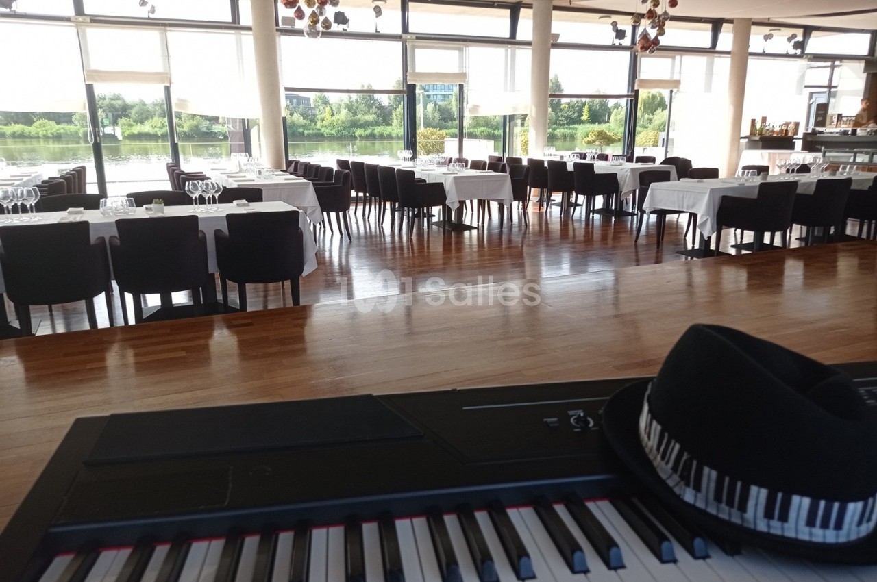 Salle de restaurant lumineuse avec des tables dressées, vue sur un piano et un chapeau posé dessus au premier plan.