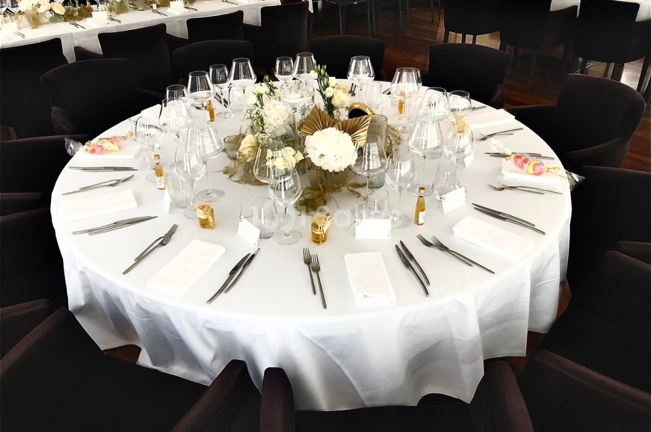 Table ronde élégamment dressée avec nappe blanche, verres, couverts, décorations florales et chaises noires.