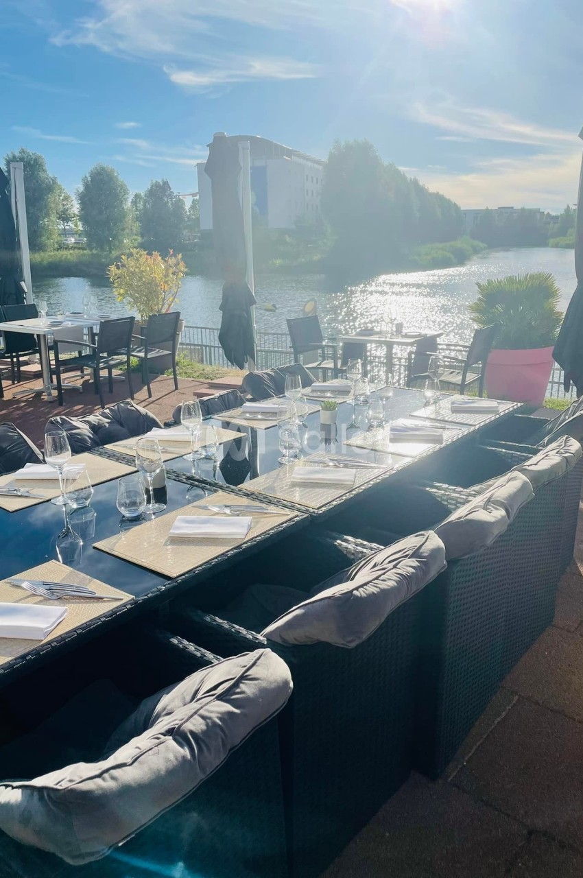 Table dressée en extérieur avec vue sur un lac, entourée de chaises en rotin et protégée par des parasols.