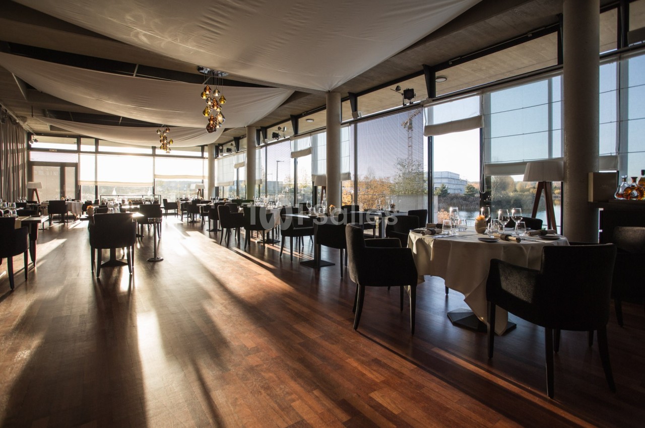 Salle de restaurant lumineuse avec de grandes baies vitrées, des tables dressées et un sol en parquet.