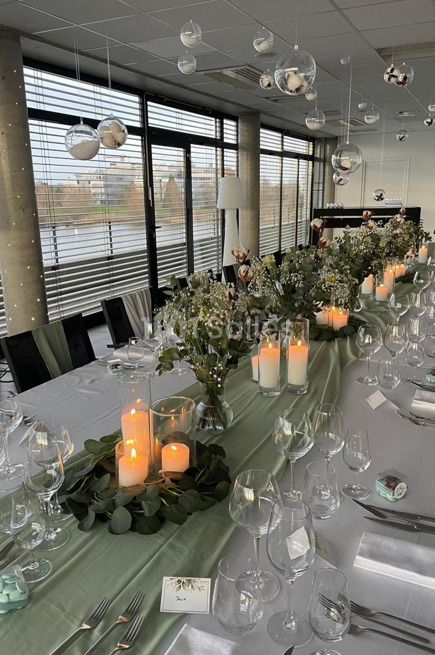 Table élégamment dressée avec nappes blanches, bougies, fleurs et décoration suspendue dans une salle lumineuse.