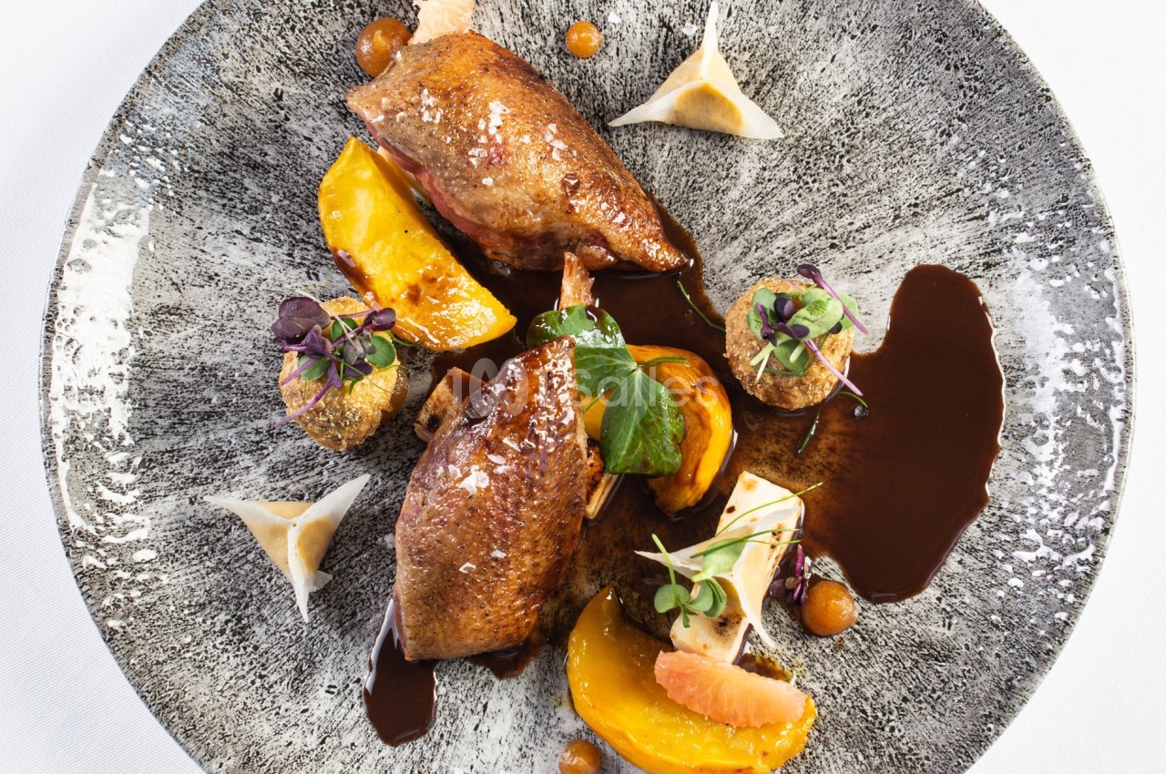 Assiette gastronomique avec viande rôtie, morceaux de fruits, sauce brune et garnitures végétales sur une assiette texturée.