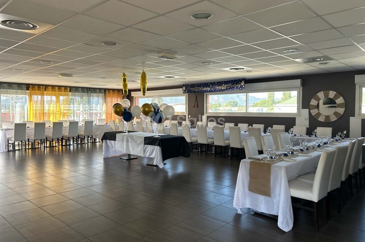 Salle de réception lumineuse avec tables dressées, chaises blanches et décorations de ballons suspendus.