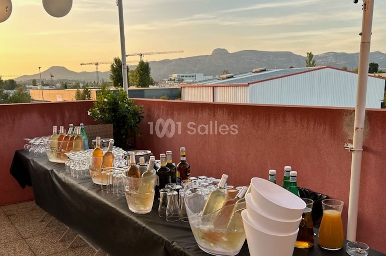 Table dressée en extérieur avec boissons, verres et seaux à glace, sur une terrasse offrant une vue sur des collines.