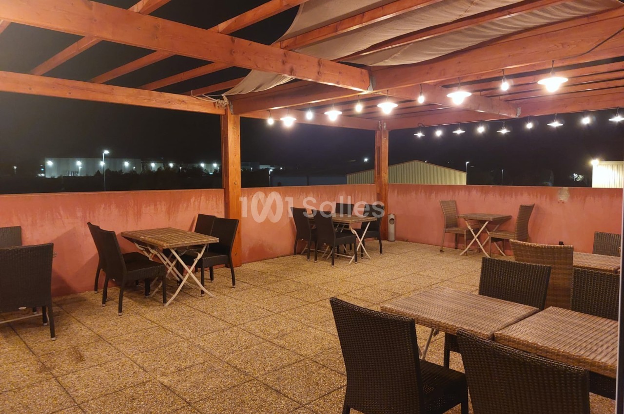 Terrasse extérieure couverte avec tables et chaises en osier, éclairée par des lumières suspendues, de nuit.