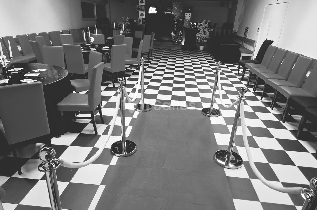 Salle avec sol à carreaux noirs et blancs, rangées de chaises, tables et cordons délimitant un tapis central.