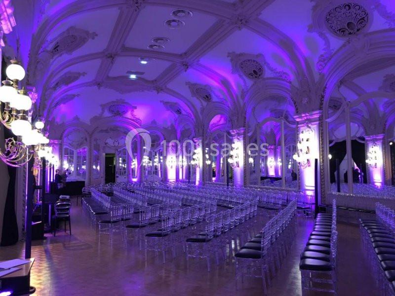 Salle de réception élégante avec plafond orné, éclairage violet et rangées de chaises transparentes alignées.