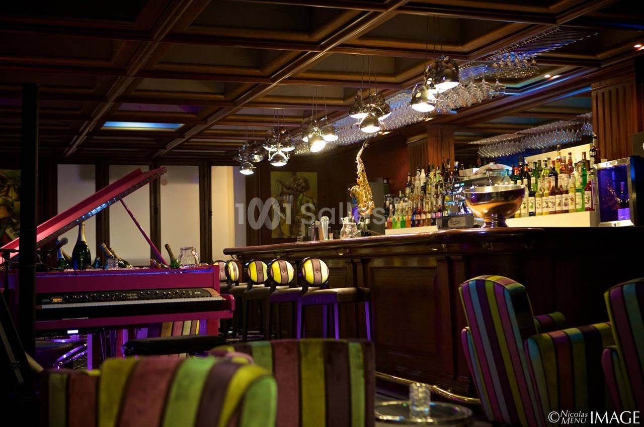 Intérieur d'un bar avec comptoir en bois, tabourets colorés, piano rose et éclairage tamisé.