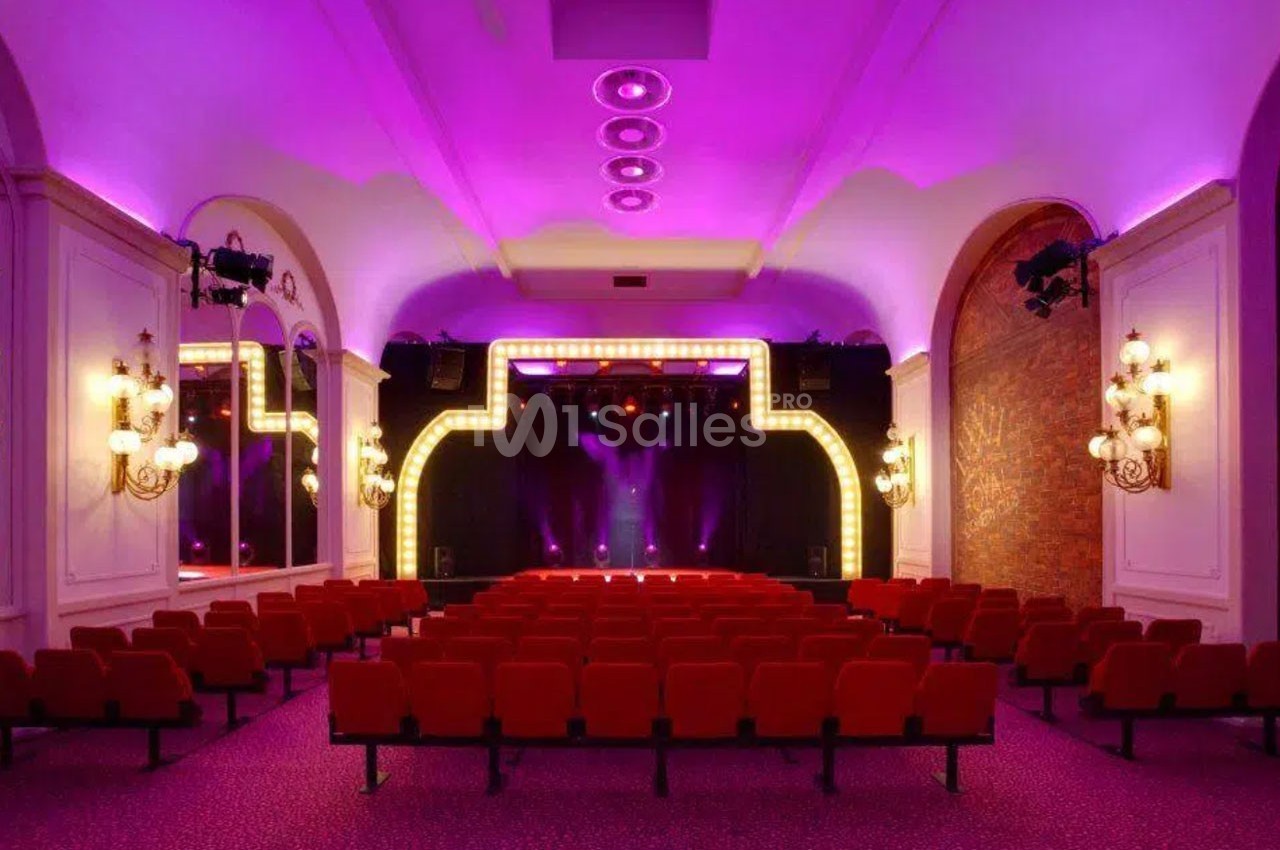 Salle de spectacle avec sièges rouges, scène éclairée par des projecteurs et ambiance lumineuse violette.