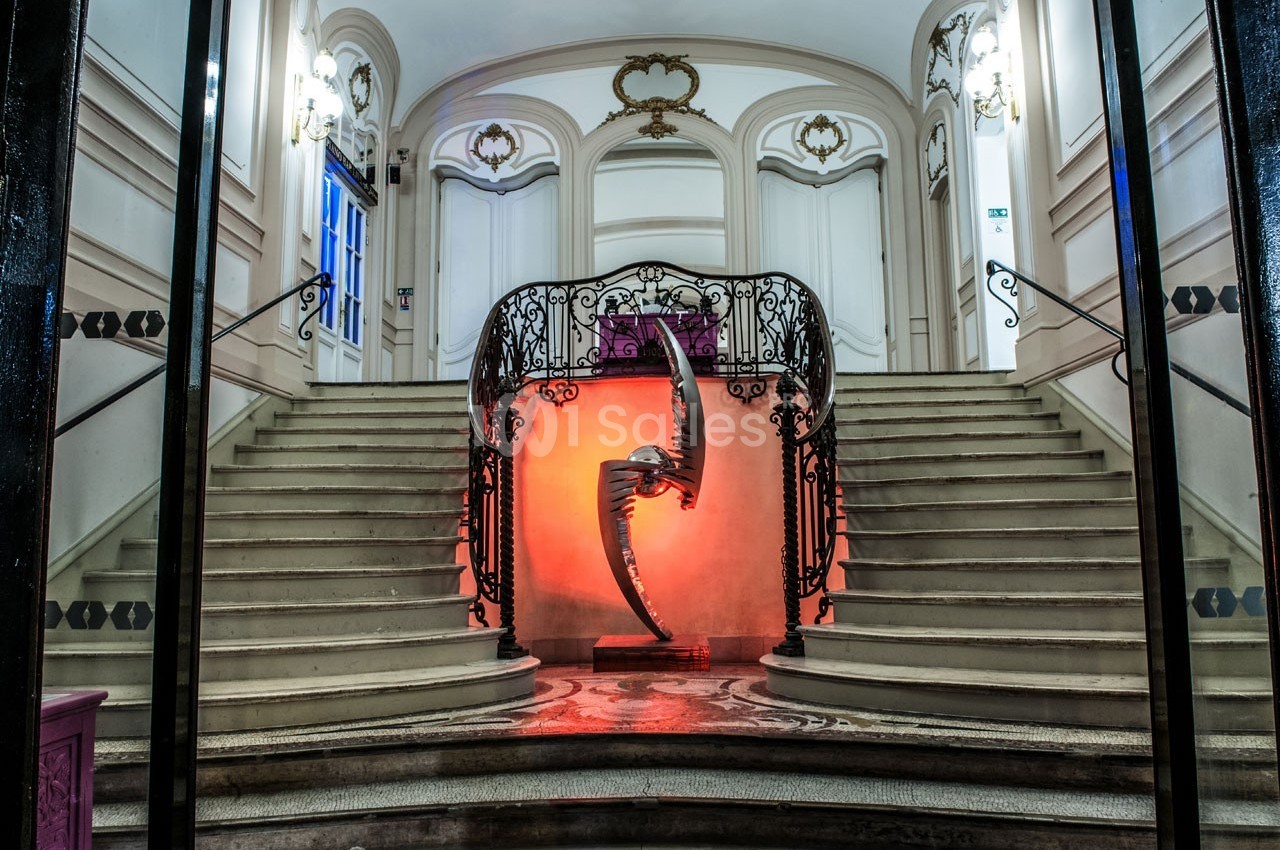 Escalier monumental avec une sculpture métallique éclairée en rouge au centre, entouré de balustrades ornées.