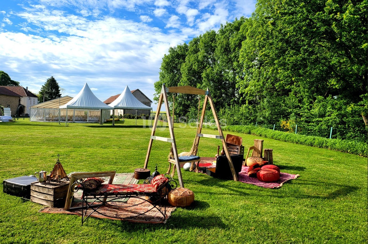 Espace extérieur aménagé avec tapis, coussins et balancelle, entouré de verdure et de tentes blanches au loin.