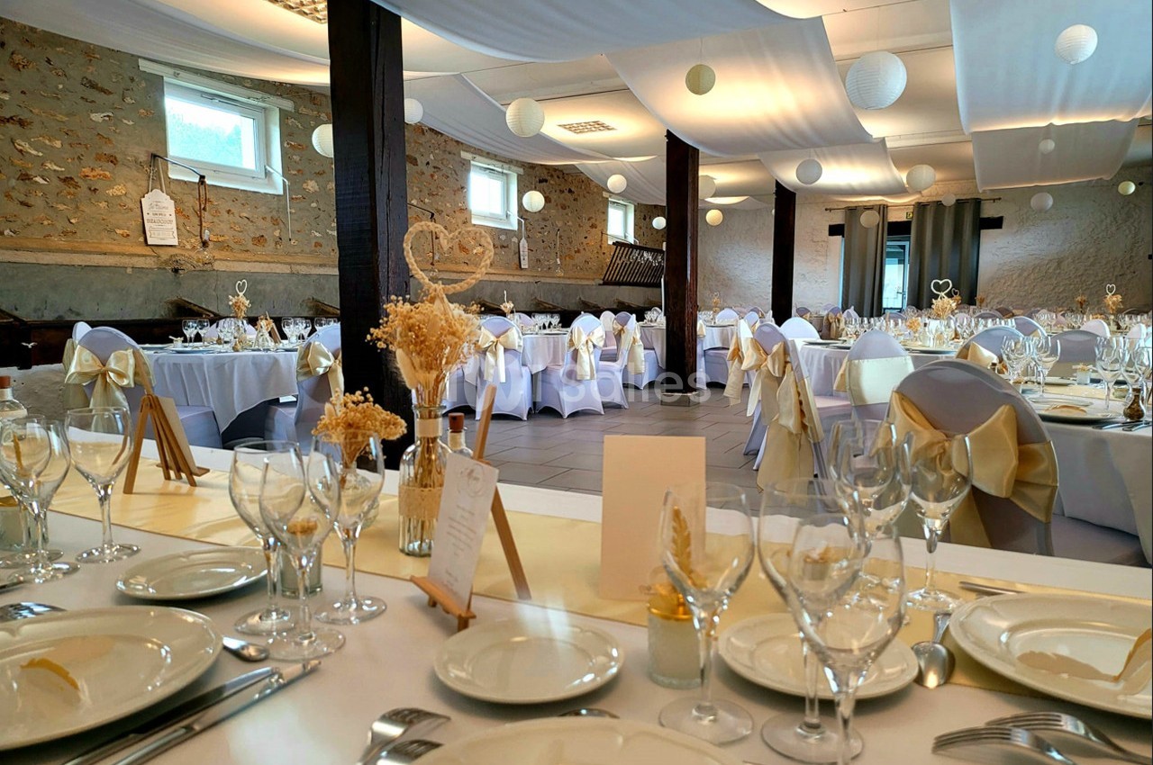 Salle de réception décorée pour un mariage, avec tables dressées, nappes blanches et chaises ornées de nœuds dorés.
