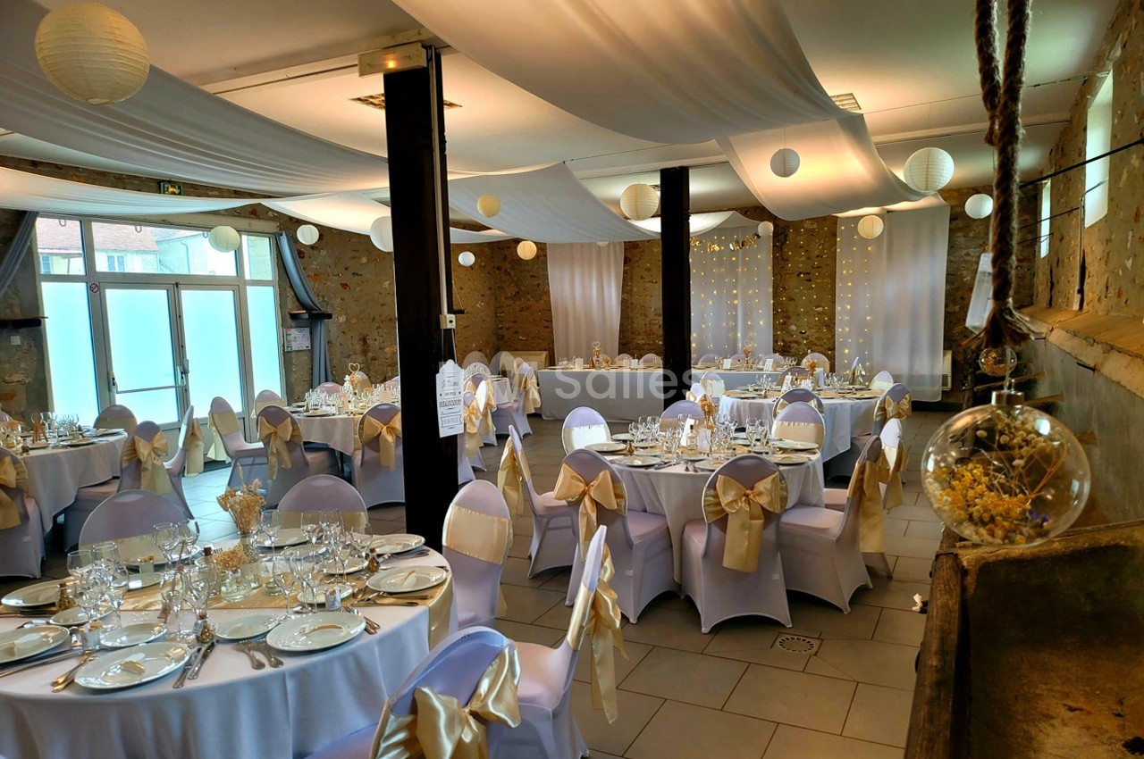 Salle de réception décorée pour un mariage, avec tables rondes dressées, nappes blanches et chaises ornées de nœuds dorés.