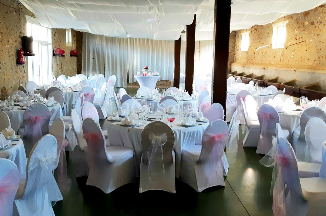 Salle de réception décorée avec des tables rondes dressées, nappes blanches et chaises couvertes de housses.