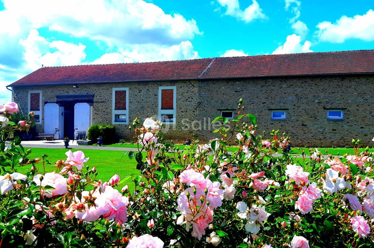 Bâtiment en pierre avec toit en tuiles rouges, entouré d'un jardin fleuri de roses sous un ciel bleu.