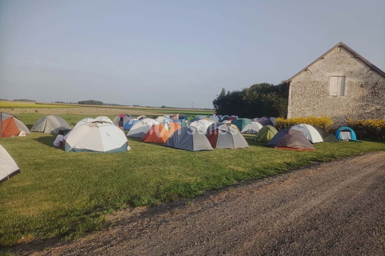 Tentes installées sur une pelouse près d'un chemin de terre et d'un bâtiment en pierre, entourées de champs.