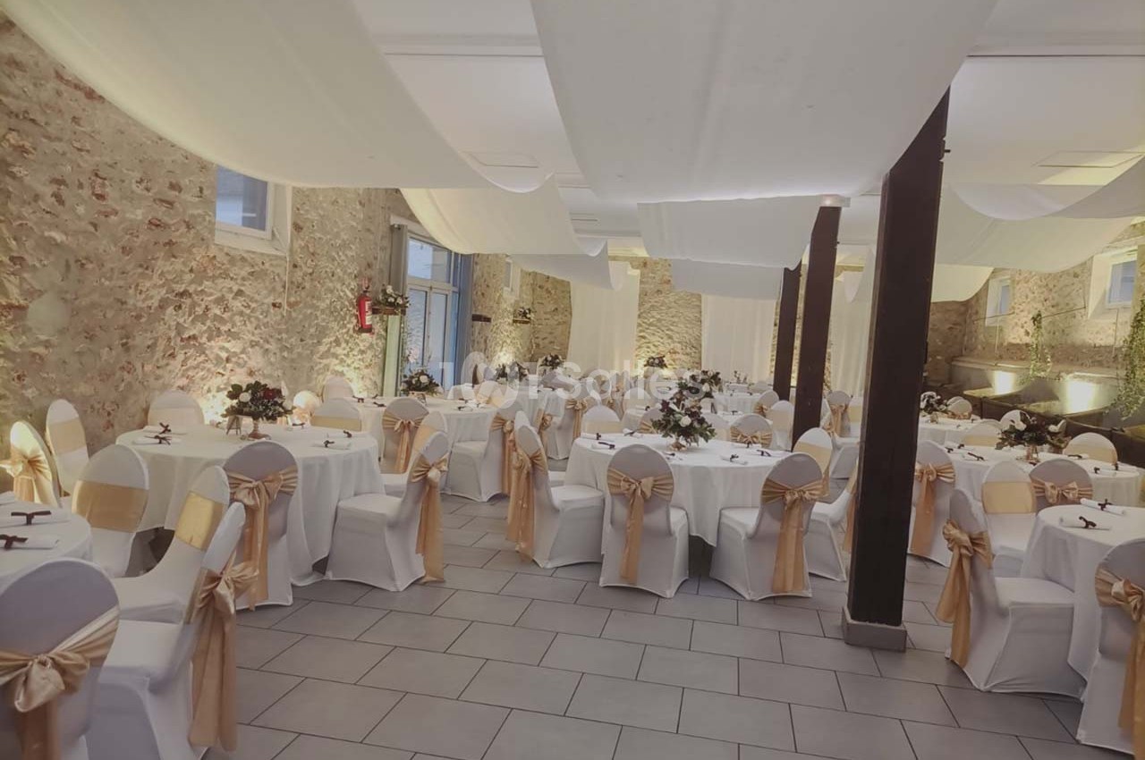 Salle de réception décorée avec des tables rondes, nappes blanches, chaises ornées de rubans dorés et murs en pierre…
