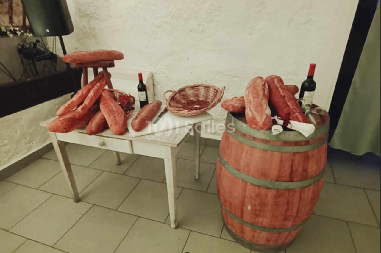 Table et tonneau avec des pains, saucissons, bouteilles de vin rouge et un panier vide dans une pièce rustique.