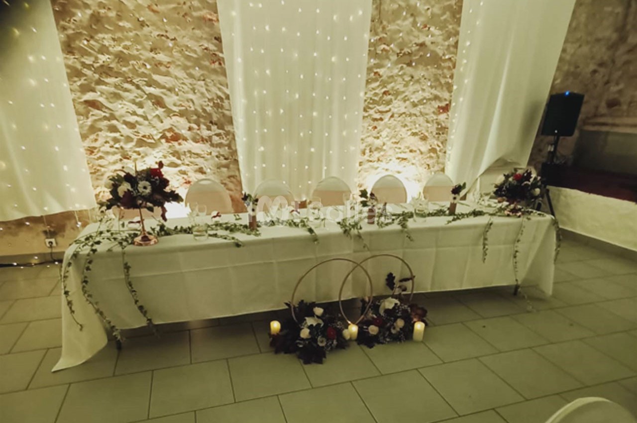 Table décorée pour un mariage avec nappes blanches, guirlandes lumineuses, fleurs et bougies devant un mur en pierre.