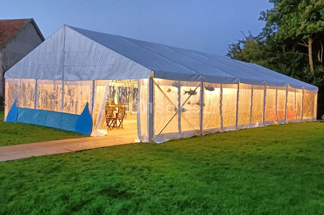 Grande tente blanche éclairée de l'intérieur, installée sur une pelouse en soirée, avec des arbres en arrière-plan.