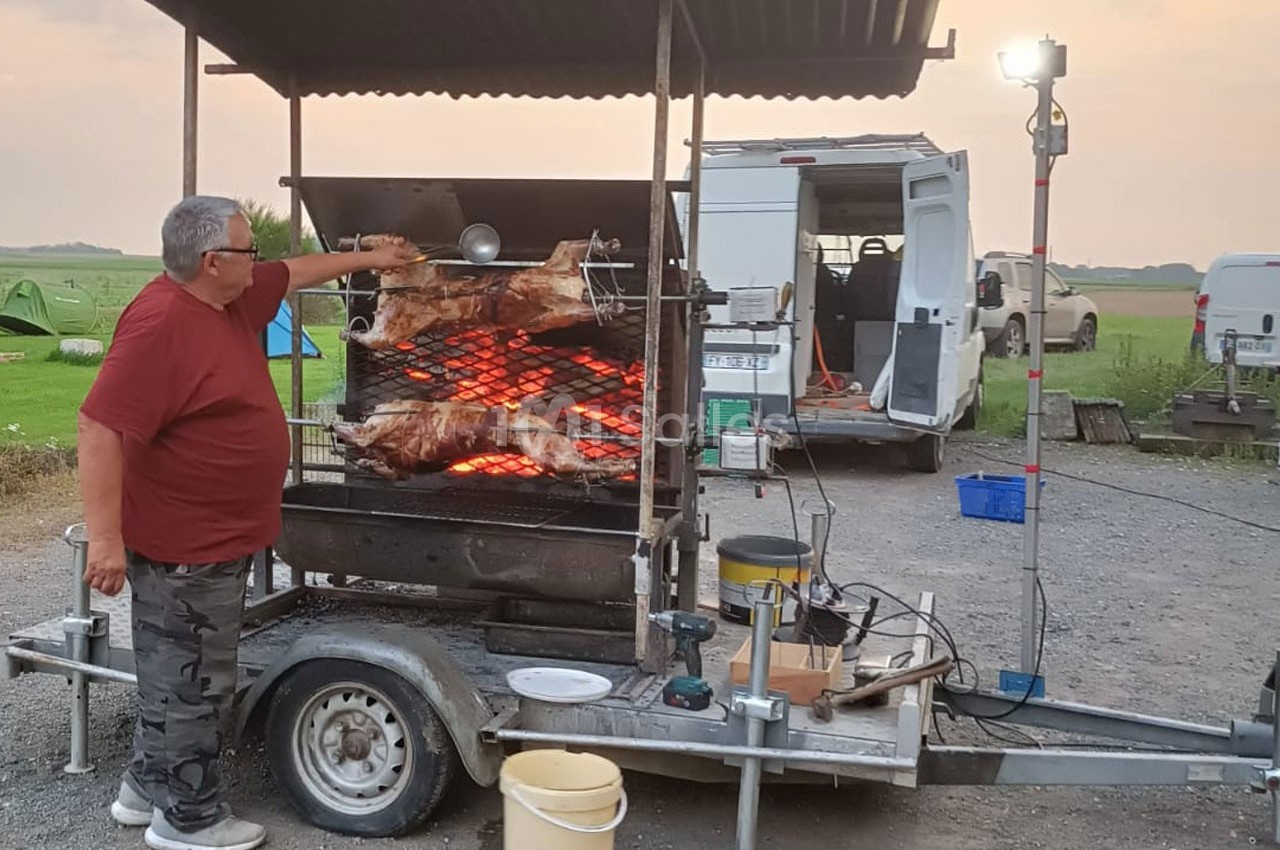 Un homme tourne des broches avec des viandes rôtissant sur un barbecue mobile en plein air.