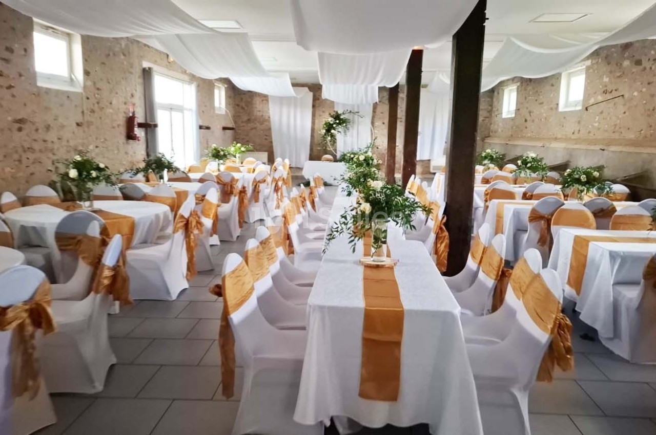 Salle de réception décorée avec des tables dressées, nappes blanches, chemins de table orange et compositions florales.